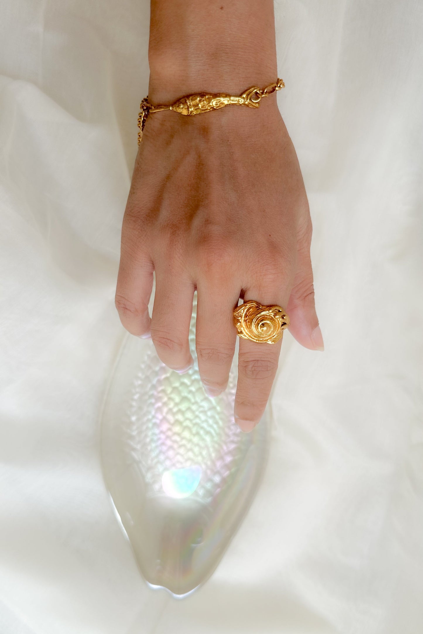 A close-up of a hand wearing a gold ring and bracelet, resting on a white textured surface next to a clear iridescent clutch bag.