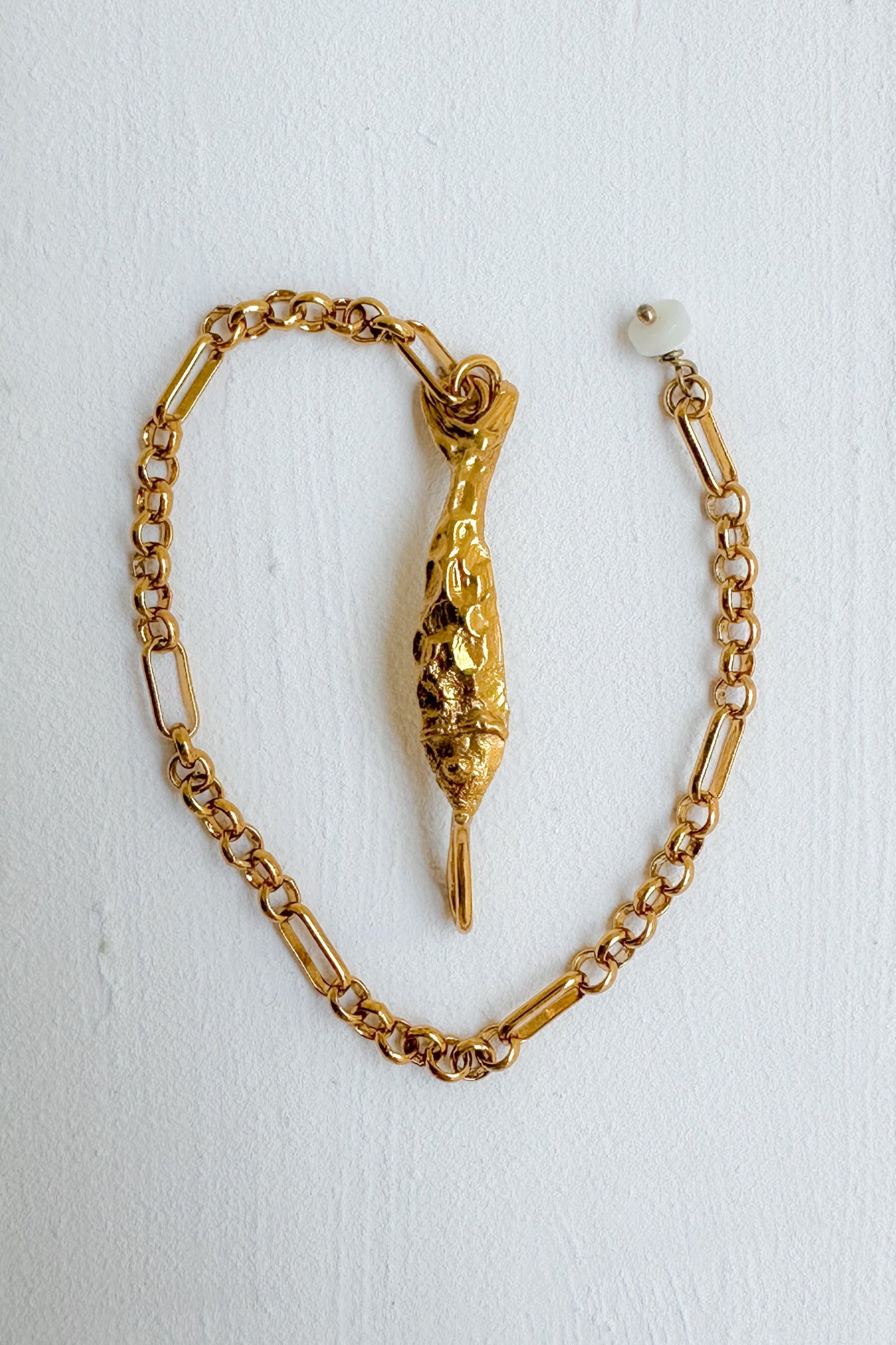 A gold chain bracelet featuring a textured, sculptural fish pendant, displayed on a white textured background.