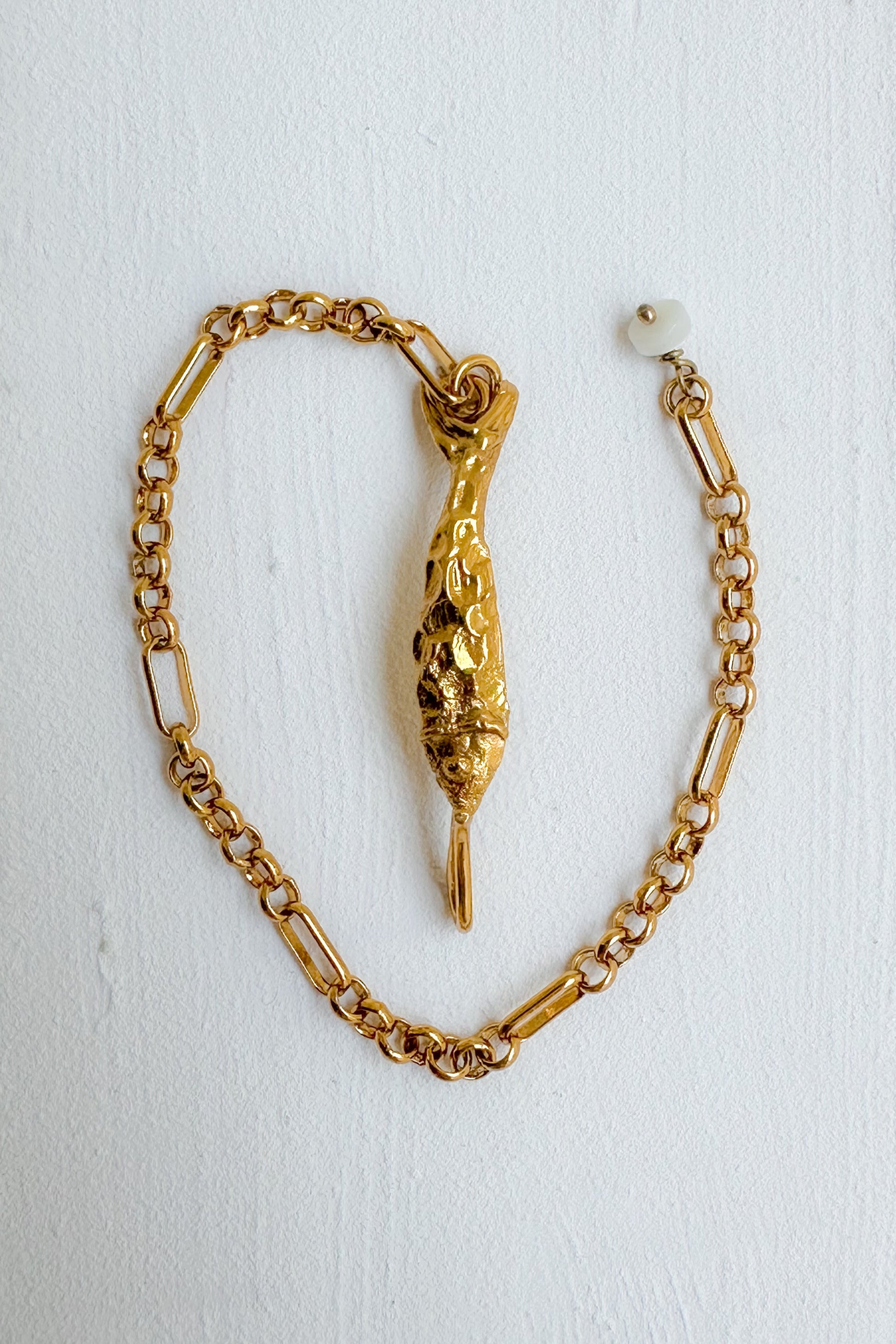 A gold chain bracelet featuring a textured, sculptural fish pendant, displayed on a white textured background.